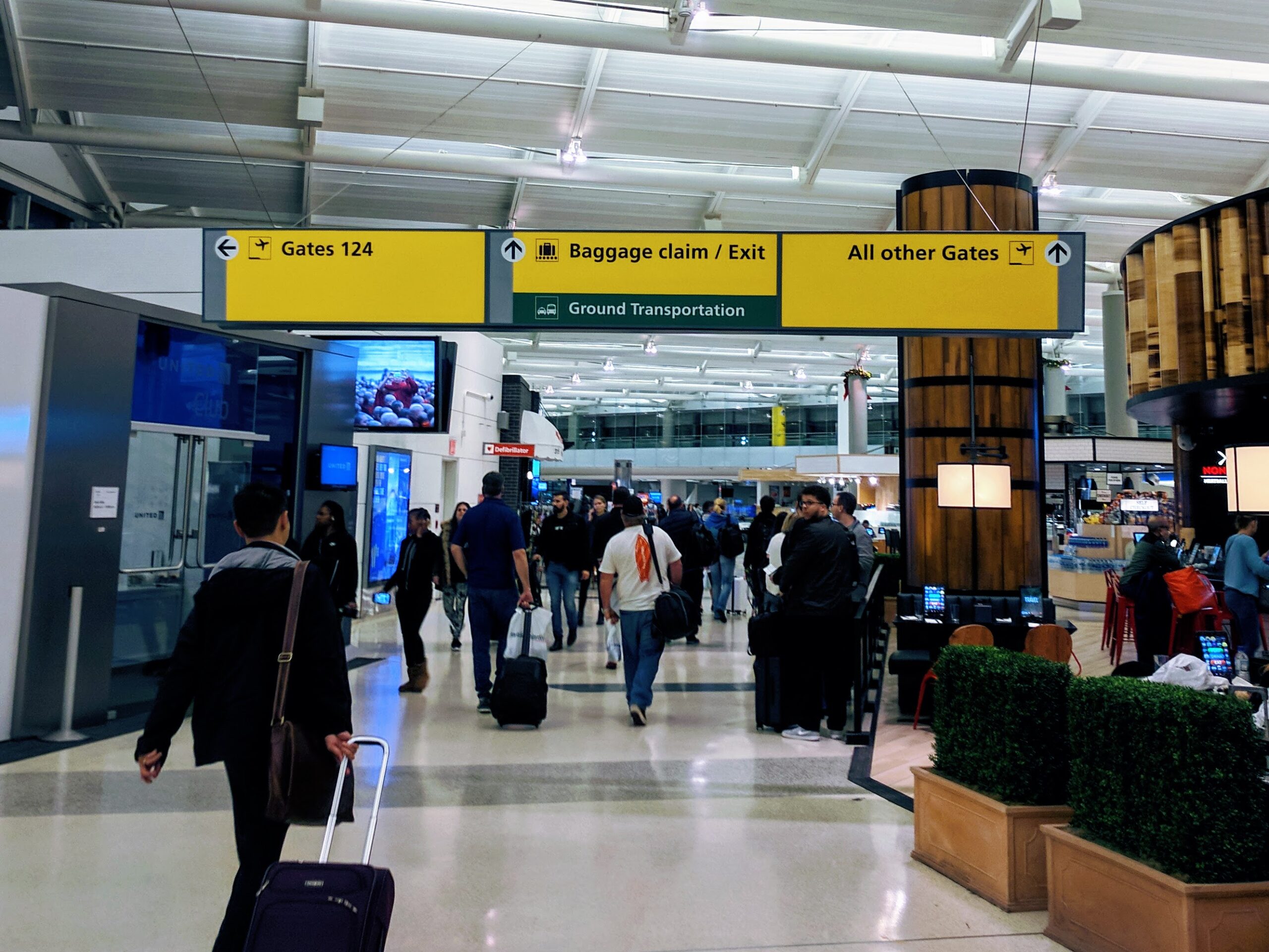 Travelers walking beneath yellow gate, baggage claim, and exit signs at Newark Liberty International Airport.
