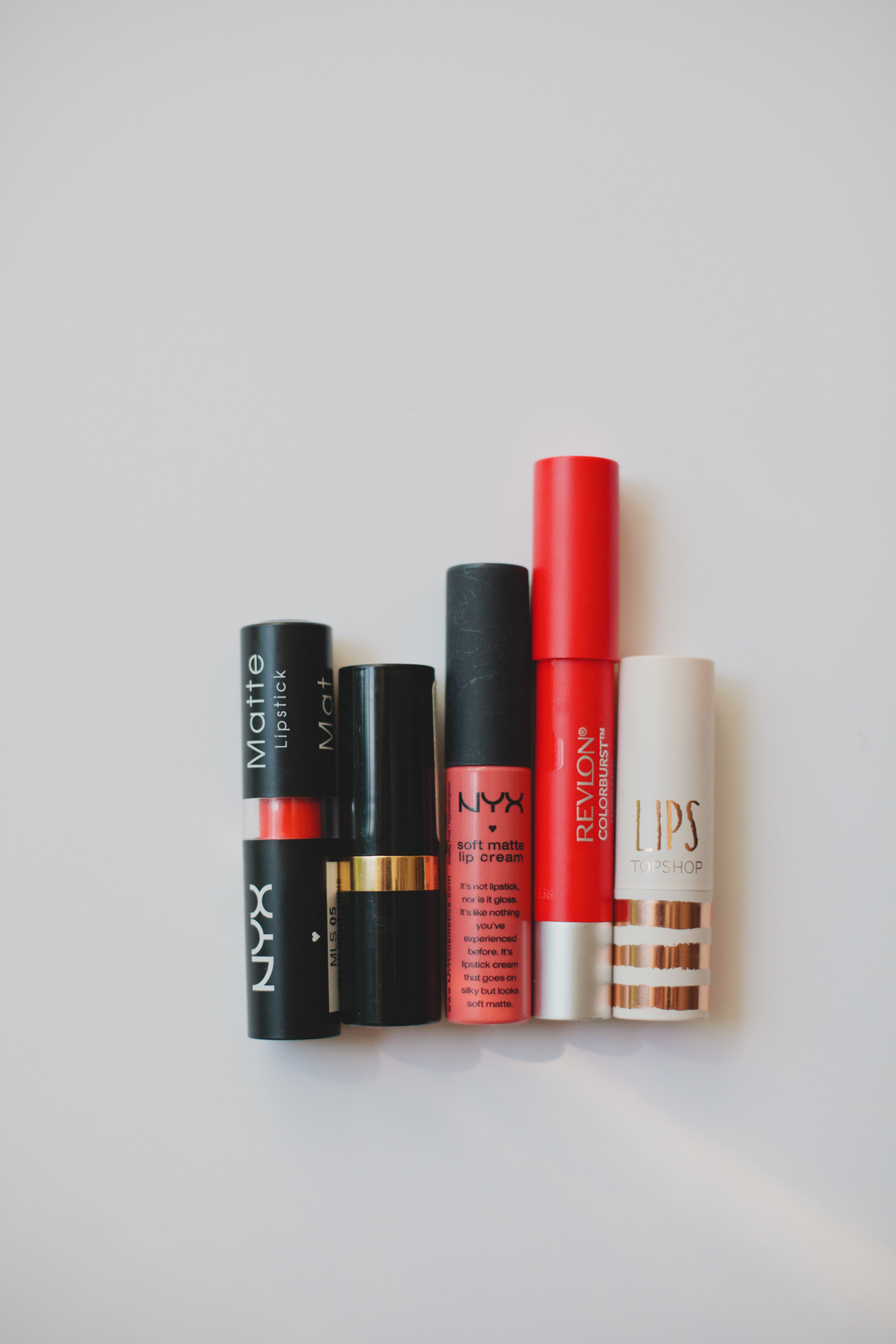 Five capped red lipstick products on a neutral surface.