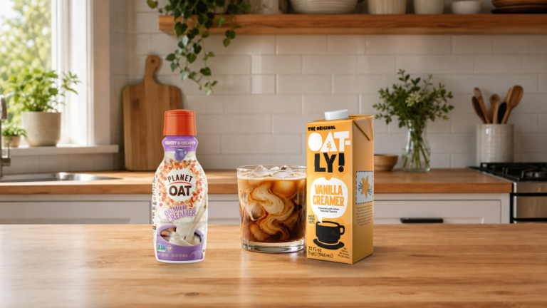 A short iced coffee glass between Planet Oat and Oatly 32-ounce vegan creamer products on a kitchen counter.