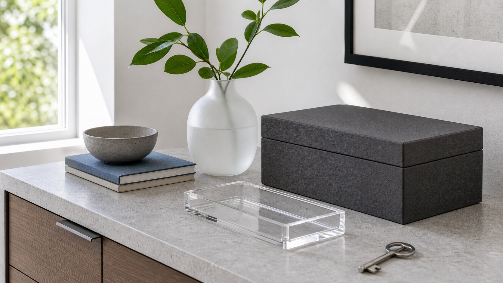 Crisp privacy still life with gray stone console, charcoal box, clear tray, white vase, green branch, and key