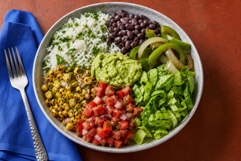 Overhead vegan burrito bowl with rice, black beans, peppers and onions, corn salsa, tomato salsa, guacamole, and lettuce.